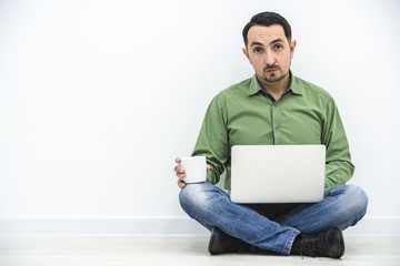 Confident and successful businessman sitting on the floor with white laptop and cup of coffeeon his knees.