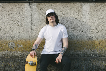 Handsome male model with beard wearing white blank t-shirt and a baseball cap with space for your logo or design in casual urban style. He is holding a skateboard