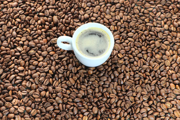 Background from coffee beans, with a white cup in the middle. Roasted coffee beans, randomly laid out on the table. Coffee drink in a white bowl.