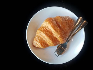 isolated plain croissant  on a white dish and utensils on black background