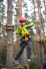 Boy climbing in an adventure park. In a green jacket and a red helmet. Overcomes obstacles.