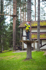 Boy climbing in an adventure park. In a green jacket and a red helmet. Overcomes obstacles.