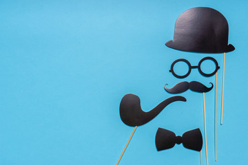 Various black photo booth props: cylinder hat, glasses, moustache, smoking pipe, bow tie on blue background. Greeting card for father's day. Creative composition in minimal style, space for text
