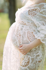 Fototapeta na wymiar Pregnant woman stands in white lace dress and supports her belly.