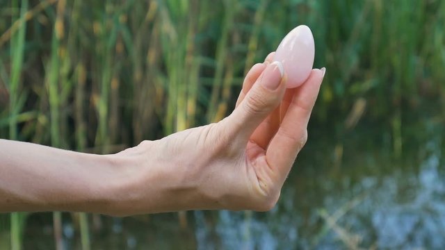 Female hand holding a rose quartz crystal yoni egg on river background. Women's health, unity with nature concepts