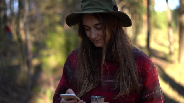 close up portrait young attractive hipster female in hat smiles and use mobile cell phone in forest camping, traveler use roaming in nature, girl looks at screen, checks social media news feed