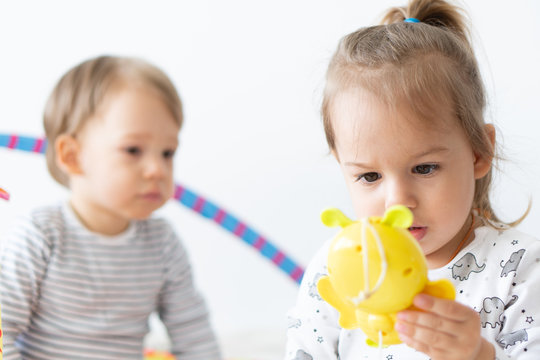 Two Small Children Play With Toys Together Sitting On The Bed. Brother And Sister Play With Colored Toys. Children With A Small Age Difference Play Together Indoors. Musical Toys That Develop Motor