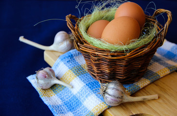 Chicken eggs. Brown chicken eggs in basket on the cutting board on blue background. Copy space. Still life with eggs, garlic, onion. Farm natural products. Egg and vegetables is ingredient for cooking