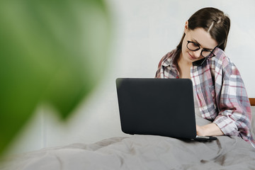 Smiling young woman wearing glasses talking on phone, sitting in bed at home with laptop, friendly manager consulting customer by smartphone, happy girl chatting with friends distracted from work