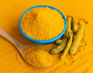 Turmeric powder in blue bowl with wooden spoon and dried roots at orange background.