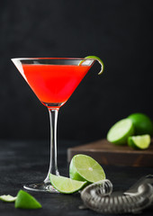 Cosmopolitan cocktail in classic crystal glass with lime peel and fresh limes with strainer on black background.