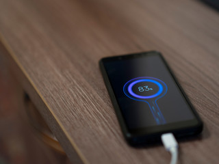 A black phone is charging at the edge of the table, a white charging cord and a blue charge indication