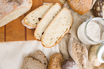 Naklejka premium Bread slices, buns and loafs on wooden table. Traditional wheat and rye bread, burlap sack, flour and white napkin. Top view. Bakery or pastry concept