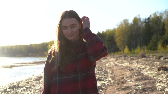 close-up front portrait of young caucasian hipster female on a wild beach with forest looking straight to camera, millennial person enjoys camping hiking holidays travel