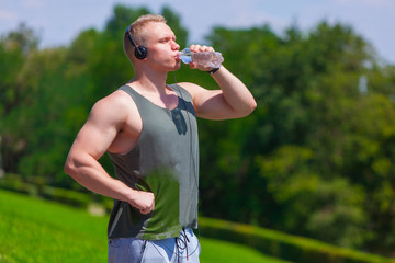 Muscular young man in headphones is drinking water and having rest after a run in summer park