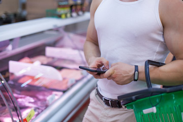 Close up of a customers shopping basket and muscular male hands holding smartphone and checking a shopping list