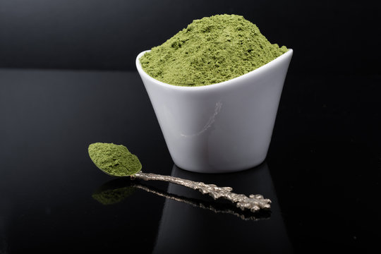 An isolated tablespoon of dried organic wheat grass and spirulina powder, on white rustic background