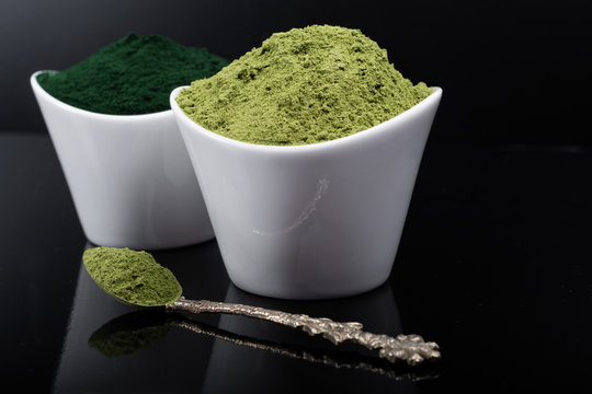 An isolated tablespoon of dried organic wheat grass and spirulina powder, on white rustic background
