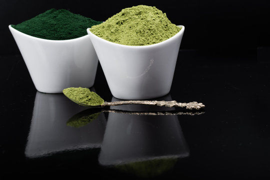 An isolated tablespoon of dried organic wheat grass and spirulina powder, on white rustic background
