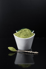 An isolated tablespoon of dried organic wheat grass and spirulina powder, on white rustic background