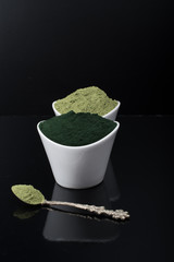 An isolated tablespoon of dried organic wheat grass and spirulina powder, on white rustic background