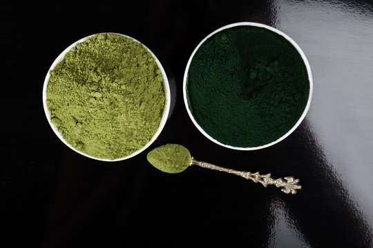 An isolated tablespoon of dried organic wheat grass and spirulina powder, on white rustic background
