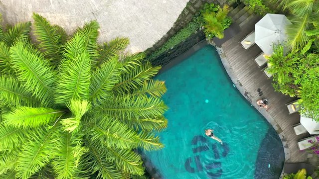 Beautiful private bungalows with reed roof and pool and around tropical palms in Bora Bora. Young woman swim in pool. Aerial view 4K