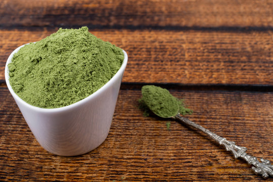 An isolated tablespoon of dried organic wheat grass and spirulina powder, on white rustic background