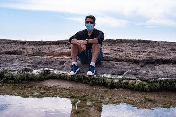 man on the beach sit on rock near a sea wearing mask for protect against infection with virus or coronavirus, civid19. enjoy sea in 2020 concept. person alone in black shirt with reflection in water
