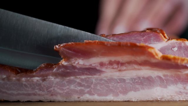 chef cuts pieces of smoked bacon by sharp knife on the wooden board