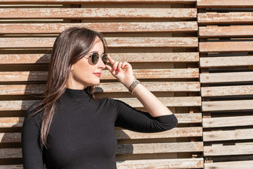 attractive long-haired girl on wooden background outdoors on a sunny day. The girl has long black hair and sunglasses. Dressed in a black sweater and casual clothes