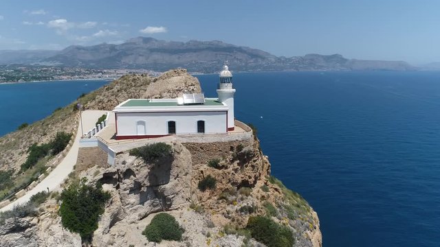 AERIAL DRONE SHOT OF MEDITERRANEAN LIGHT HOUSE IN A CLIFF WITH COAST AND WHITE VILLAGE IN THE BACKGROUND