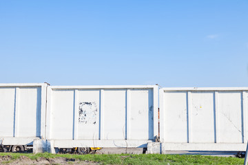 A concrete fence of white color with green grass at the base, enclosing the territory of an industrial enterprise on which special equipment stands against a blue sky.