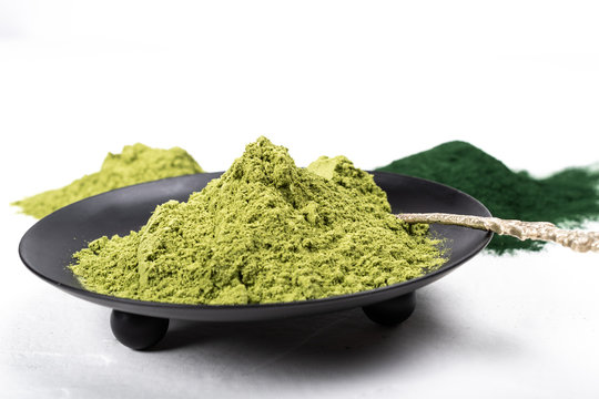 An isolated tablespoon of dried organic wheat grass and spirulina powder, on white rustic background