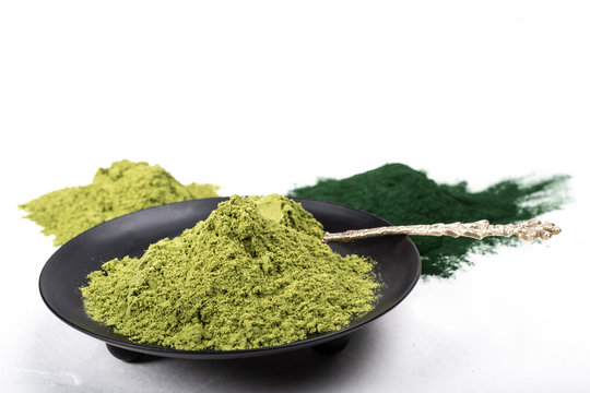 An isolated tablespoon of dried organic wheat grass and spirulina powder, on white rustic background