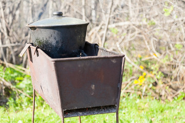 Black smoked cast-iron cauldron with a lid on coals in a metal brazier against a background of green grass. Cooking pilaf with handy means on an open fire in nature.