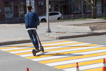 A young guy in a blue bomber jacket and jeans on an electric scooter moves very quickly along a pedestrian crossing, leaning to his side. Modern environmental gadget.