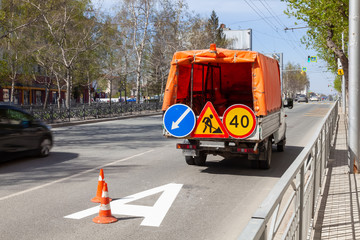 Work car with orange tarpaulin and traffic signs indicating the direction of detour warning about road work next to the bus stop markings and road cones.