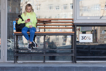 Obraz na płótnie Canvas A blonde girl in a bright yellow sweatshirt sits on a wooden bench with a backpack near a coffee shop, holds a phone in her hands and waits for her order. The girl at the bus stop.