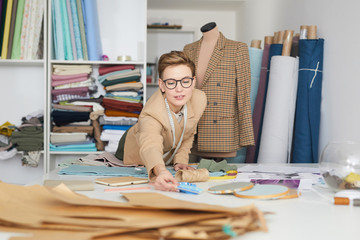 Young tailor in eyeglasses working at the table with fabrics in the workshop she making pattern for future clothes