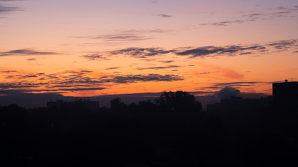 Fototapeta premium Sunrise over the dark city. Dark silhouettes of residential buildings and trees against a bright, orange sky with clouds.