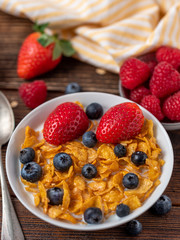 Corn flakes with blueberries and raspberries in white bowl in dark wooden desk.