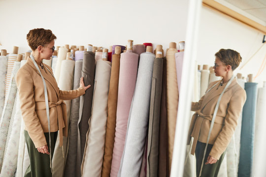 Young designer standing near the rolls with textile and choosing the fabric for future clothes in workshop