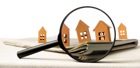 Magnifier in front of an open newspaper with paper houses on white background. Concept of rent,...