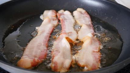 Bacon sizzling in a frying pan. Delicious, unhealthy breakfast meat fries in a skillet. Pork products are experiencing a supply chain issue and shortages due to the Coronavirus pandemic.
