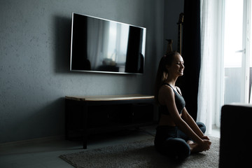 A girl appearance sits on the floor in a butterfly pose. He trains at home near the TV next to the window. Self isolation athlete