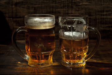 A glass of amber beer. Glasses on a wooden table. Black shabby boards on the background. Bright contrasting light and glare on the glass.