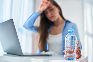Upset tired working woman suffering from heat, thirst and hot weather cools down with cold water bottle during online work at computer at warm summer day - obrazy, fototapety, plakaty