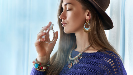Stylish fashionable attractive brunette boho chic woman with closed eyes wearing jewelry and hat holds perfume bottle - obrazy, fototapety, plakaty