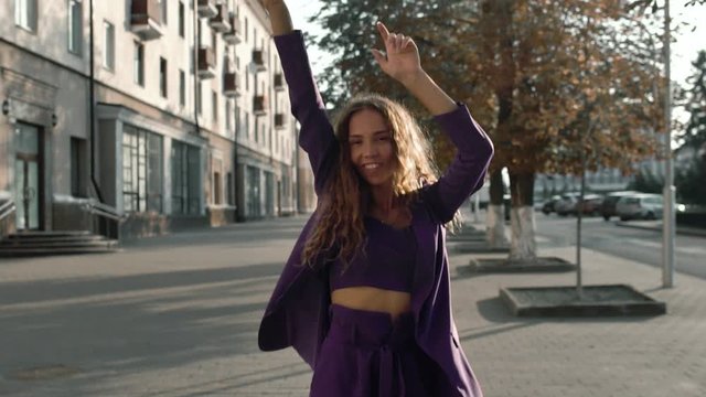 Happy curly long haired smiling girl dancing outdoors at city street. Fashionable stylish woman in suit moves in slow motion. Positive concept. Vogue female looks into a camera. Trendy business lady